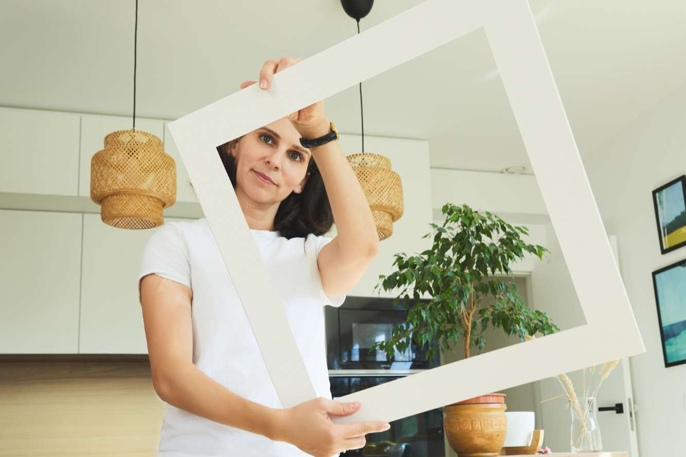 Woman holding a passepartout in a room with a plant and light fixture.