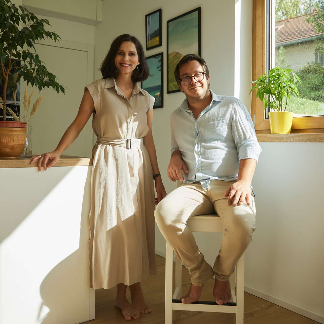 Two people in a bright room with plants and artwork on the wall.